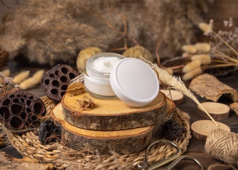 Opened glass cream jar with blank lid on wood near natural decorations close up, package mockup. Eco friendly skincare product, lotion or cream. Bohemian composition with dried leaves and flowers.