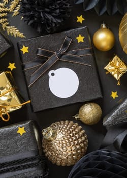 Black and golden Christmas Gift Boxes with a bow and ornaments close up, round paper gift tag mockup, copy space. Dark winter composition with blank label card for Christmas, New Year, Birthday, Anniversary