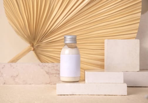 Small bottle with blank label on travertine stone against dry beige palm leaf close up, mockup. Skincare beauty product - cream or lotion. Natural organic cosmetics. Minimal eco friendly composition..