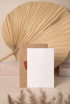 Blank card near beige travertine stones against dry beige palm leaf close up, mockup. Boho scene with vertical paper card. Bohemian or close to nature greeting or wedding Invitation. Minimal composition.