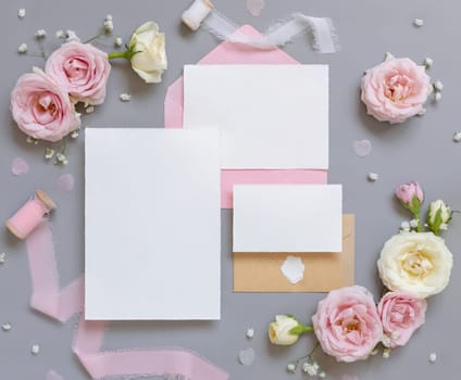 Paper cards and envelopes between light pink roses and silk ribbons on grey top view, wedding mockup. Romantic scene with blank cards and pastel flowers. Valentines, Spring or Mothers day concept