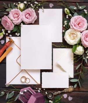 Cards and envelope between pink and cream roses on brown wood top view, wedding mockup. Romantic scene with cards, envelope, rings and pastel flowers flat lay. Valentines, Spring or Mothers day concept