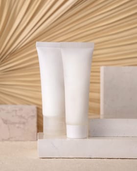 White blank tubes on travertine stone against dry beige palm leaf close up, mockup. Skincare beauty product - cream or lotion. Natural organic cosmetics. Minimal eco friendly composition..