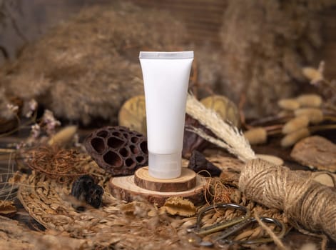White plastic cosmetic tube on wood near natural decorations closeup, packaging mockup. Eco friendly skincare product, lotion or cream. Bohemian composition with dried leaves and flowers.