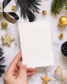 Hand holding Card near black and golden Christmas decorations and fir tree branches, top view. Winter flat lay with blank invitation card, Mockup, copy space