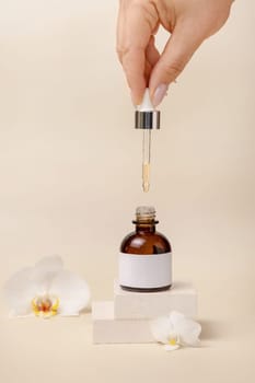 Hand holding pipette over opened cosmetic dropper bottle near white orchid flowers and stones on light beige, close up, mockup. Natural skincare product, serum or essential oil.