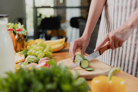 Close up on young housewife woman or girl hands slicing cucumber with kitchen knife, cuts food on wooden chopping board. Unrecognizable female standing at kitchen counter, cutting organic vegetables