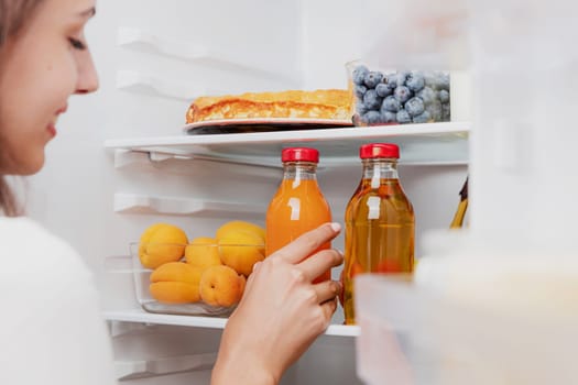 Woman hand taking, grabbing or picks up juice glass bottle out of open refrigerator shelf or fridge drawer full of fruits, vegetables, banana, peaches, yogurt. Healthy food diet, lifestyle concept