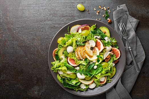 Fresh green leafy salad with mozzarella cheese, fruits and nuts on dark background close up. Concept for a tasty and healthy meal