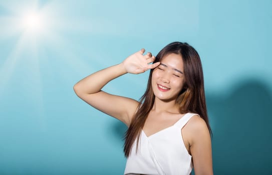 Asian young woman hand up cover face protect sun light and UV smile in studio shot isolated on blue background, female raising arm and hand in protection and block sunshine, sunscreen cream advertise