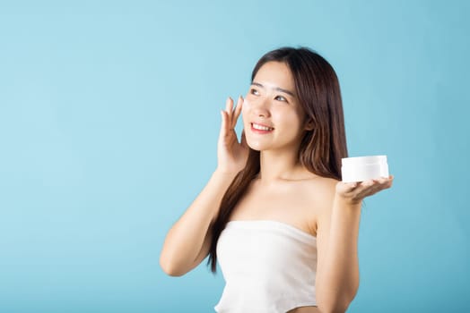 Asian young woman are not very beautiful hold jar of skin cream for face and apply anti-aging moisturizer studio isolated on blue background, Happy female pointing finger her face, pursuit of beauty