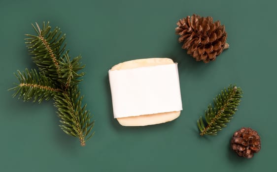 Soap bar with blank label near fir tree branches and pine cones on green top view. Holiday brand packaging mockup. Skincare beauty product, cream or serum. Healthcare natural cosmetics