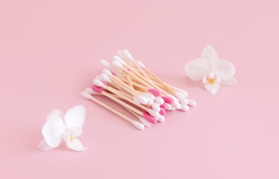 White and pink bamboo cotton buds near white orchid flowers on light pink, close up. Organic skincare product for everyday beauty routine. Eco friendly Biodegradable cotton swabs..