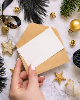 Hand holding Card and envelope, black and golden Christmas decorations and fir tree branches, top view. Winter flat lay with blank invitation card, Mockup, copy space