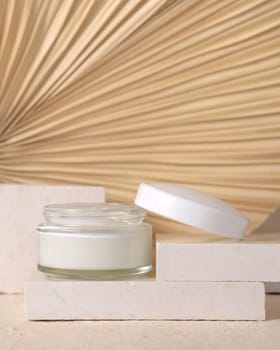 Opened glass cream jar with a white lid on travertine stone against dry beige palm leaf close up, mockup. Skincare beauty product - cream or lotion. Natural organic cosmetics. Eco friendly composition..