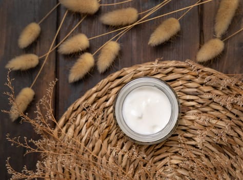 Opened cream jar on wattled placemat on wood near Hare's Tail grass top view. Natural skincare product for everyday beauty routine. Bohemian eco friendly cosmetics flat lay.