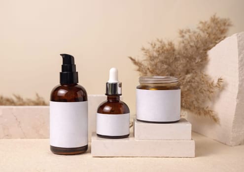Glass bottles on travertine stone against dry pampas grass on beige close up, mockup. Skincare beauty products - cream, essence or lotion. Natural organic cosmetics. Minimal eco friendly composition..