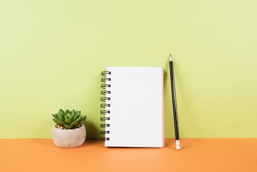 Blank notepad with a pencil on an orange and green background, pot with a cactus, copy space for text, office desk
