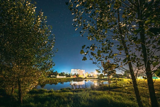 Night Sky Over River Lake Near Residential Houses. Night Starry Sky Above Lake River With Bright Stars. Glowing Stars Above Summer Nature. Photo Among Trees.
