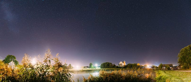 Panorama Of Evaporation Over River Lake Near Houses In Village. Night Starry Sky Above Lake River With Bright Stars And Meteoric Track Trail. Glowing Stars Above Summer Nature.