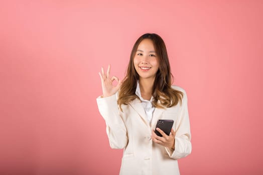 Happy Asian portrait beautiful cute young woman excited holding mobile phone and gesturing ok sign for agree, studio shot isolated on pink background, Thai female making finger symbol on smartphone