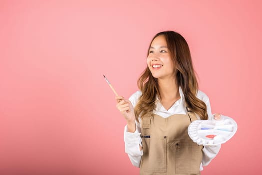 Happy female painting using paintbrush and palette with colors, Asian beautiful young woman artist holding brush and paint palette, studio shot isolated on pink background, Paintings and art equipment
