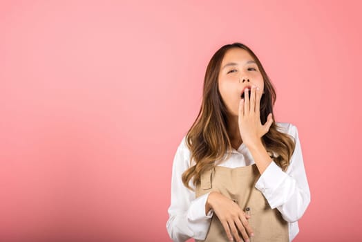 Portrait Asian beautiful young woman wearing overalls emotions tired and sleepy her yawning covering mouth open by hand, studio shot isolated on pink background, Female attractive insomnia boring