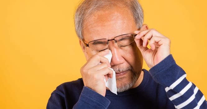 Portrait Asian senior old man sad wiping away his tears studio shot isolated on yellow background, Elder man crying raise glasses with tissue wipe red eyes, Sadness depressed lonely