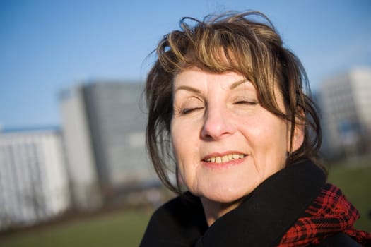 Side head portrait of a mature woman against a fuzzy urban background with closed eyes deeply breathing