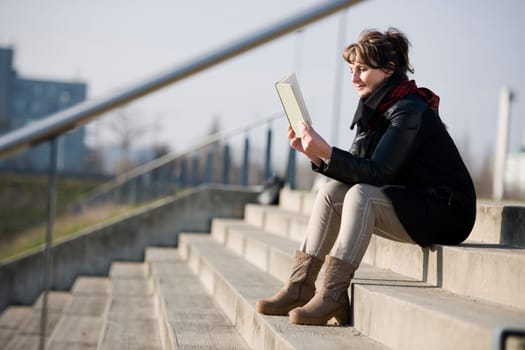 Side full body view of a mature woman in black leather jacket on a concrete staircase in a book reading against a fuzzy urban background