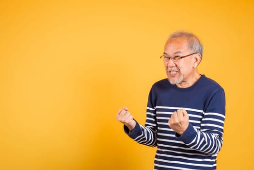 Portrait smiling senior old man lifestyle he arms raised to celebrating his success, Happy Asian elder man with glasses excited say yes gesture like winner studio shot isolated on yellow background