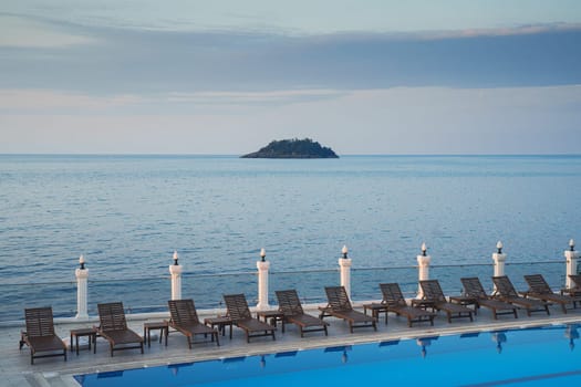 wooden sunlounger chairs near a swimming pool with blue water with sea and a small island in the background at sunset