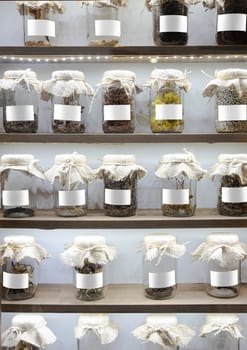 front view closeup of hand made glass jar recipients containing dried spices and ingredient condiments with empty white labels and white fabric enclosures stacked on pantry shelves