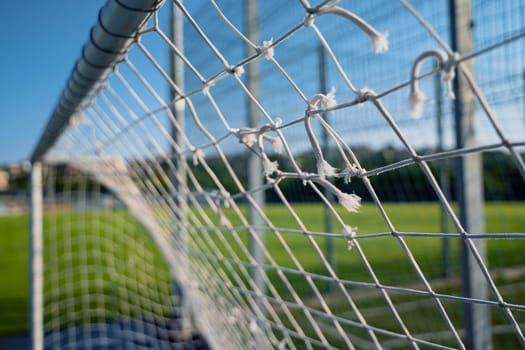 closeup of damaged soccer gate goalpost white rope net with knots and green field and blue sky in background