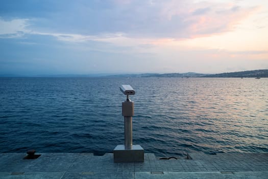 rear view closeup of coin operated metallic binoculars on concrete pier at the sea shore with sunset sky in the background