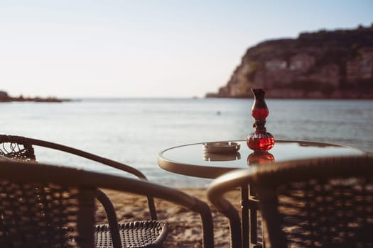 rear view closeup of empty cafe table and chairs on sandy beach with ashtray and candle light in front of sea golf with clear sky before sunset
