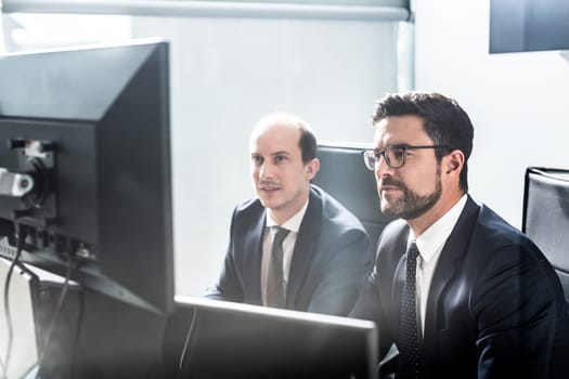 Image of two thoughtful businessmen looking at data on multiple computer screens, solving business issue at business meeting in modern corporate office. Business success concept.