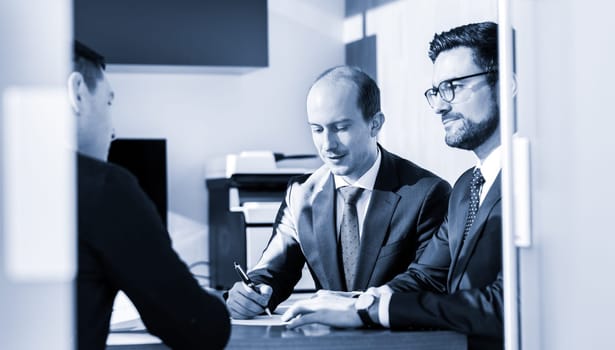 Team of confident successful business people reviewing and signing a contract to seal the deal at business meeting in modern corporate office. Business concept. Greyscale blue toned image.