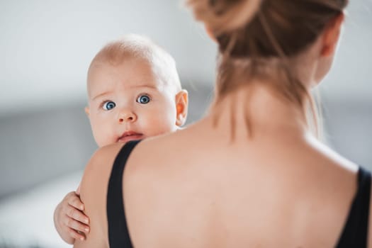 Portrait of sweet baby resting in mothers arms, looking at camera, touching mama shoulder. New mom holding little kid, embracing child with tenderness, love, care. Motherhood concept.