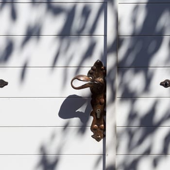 Traditional mediterranean background consisting of arhitectural detail of white wooden door with brass doorknob with olive tree shadow