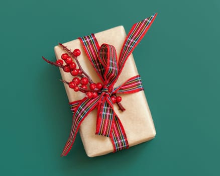 Gift box wrapped in karft paper with red ribbon bow and berries on green top view. Christmas or New Year Holiday atmosphere flat lay