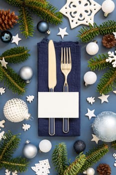 Blue and white Christmas table setting with blank card near decorations and fir tree branches top view. Winter table with fork, knife and plate. Winter New Year or Christmas dinner atmosphere