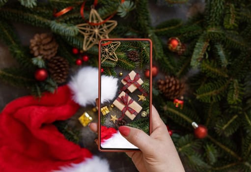 Woman's hand taking a mobile photo of christmas composition with Santa Claus hat, fir branches and holiday decorations top view. Blogger preparing content for social media using cell phone.