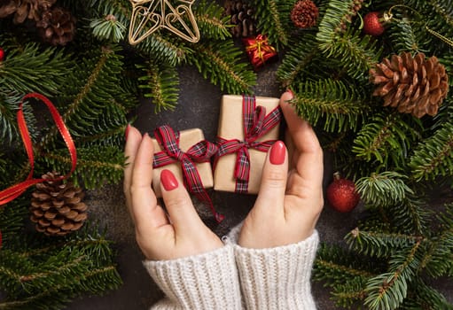 Female hands holding gift boxes near Christmas decorations and fir tree branches top view on a dark table. Christmas holiday atmosphere flat lay. Wrapping presents for loved ones during winter holidays