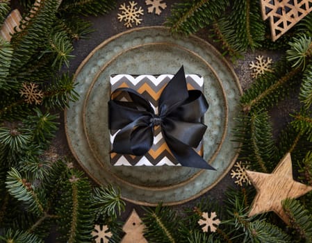 Wrapped present on Festive table near fir tree branches and Rustic Christmas decorations top view. Christmas or New Year table place with gift box on green plates on dark table flat lay