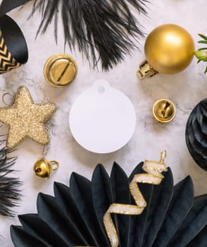 Paper round gift tag between fir tree branches and black and golden Christmas decorations on a white marble table top view. Mockup, copy space. Winter holiday flat lay with blank label