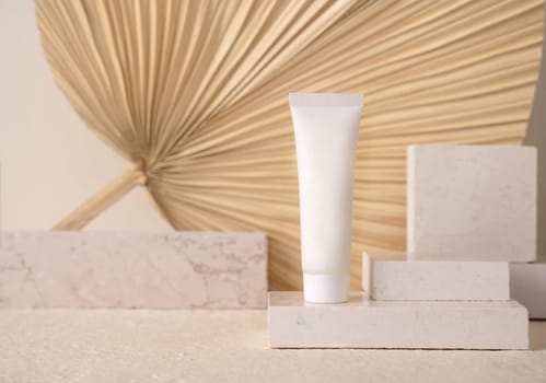 White blank tube on travertine stone against dry beige palm leaf close up, mockup. Skincare beauty product - cream or lotion. Natural organic cosmetics. Minimal eco friendly composition..