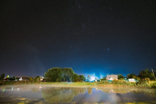 Evaporation Over River Lake Near Houses In Village. Milky Way Galaxy In Night Starry Sky Above Lake River Landscape At Night. Glowing Stars Above Summer Nature