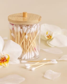 Bamboo cotton buds in a jar near white orchid flowers on light beige, close up. Organic skincare product for everyday beauty routine. Eco friendly Biodegradable cotton swabs..