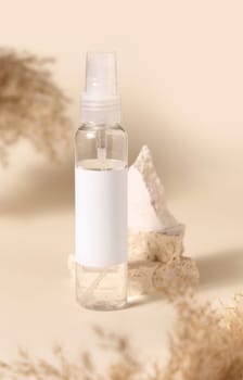 Transparent glass cosmetic bottle with blank label on stones near pampas grass on light yellow, close up, mockup. Skincare beauty product. Natural eco friendly cosmetics
